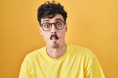 Hispanic man wearing glasses standing over yellow background making fish face with lips, crazy and comical gesture. funny expression. 