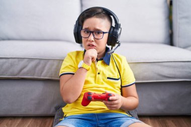 Young hispanic kid playing video game holding controller wearing headphones thinking worried about a question, concerned and nervous with hand on chin 
