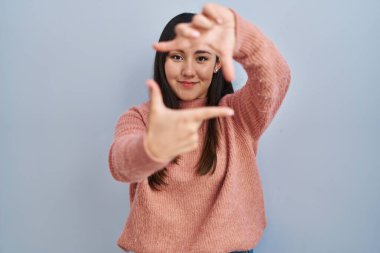 Young latin woman standing over blue background smiling making frame with hands and fingers with happy face. creativity and photography concept. 