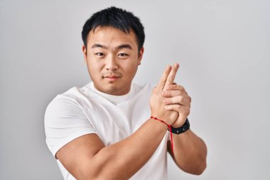 Young chinese man standing over white background holding symbolic gun with hand gesture, playing killing shooting weapons, angry face 