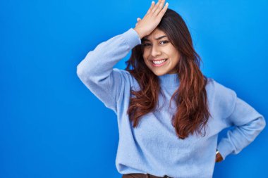 Hispanic young woman standing over blue background surprised with hand on head for mistake, remember error. forgot, bad memory concept. 