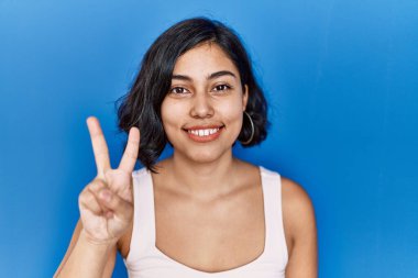 Mavi arka planda dikilen genç İspanyol kadın iki numaralı parmaklarını gösterip işaret ederken kendinden emin ve mutlu gülümsüyor.. 