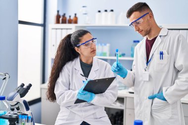 Man and woman wearing scientist uniform write on clipboard holding test tube at laboratory