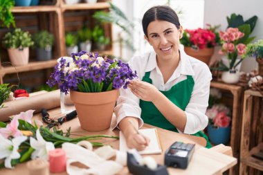 Young beautiful hispanic woman florist writing on notebook touching plant at flower shop