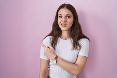 Young hispanic girl standing over pink background pointing aside worried and nervous with forefinger, concerned and surprised expression 