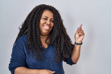 Plus size hispanic woman standing over white background with a big smile on face, pointing with hand and finger to the side looking at the camera. 