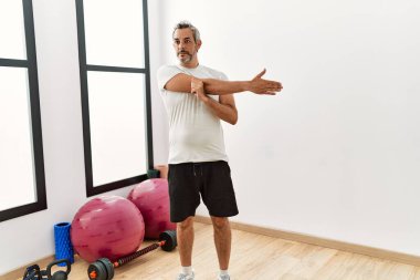 Orta yaşlı, kır saçlı bir adam spor merkezinde kolunu esnetiyor.