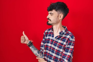 Young hispanic man with beard standing over red background looking proud, smiling doing thumbs up gesture to the side 