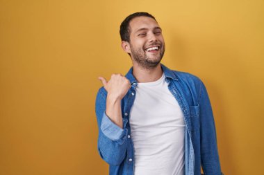 Sarı arka planda dikilen İspanyol adam gülümsüyor, gülümsüyor ve başparmağıyla yan tarafa bakıyor.. 