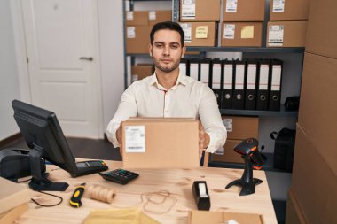Young hispanic man with beard working at small business ecommerce holding box relaxed with serious expression on face. simple and natural looking at the camera. 