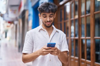 Genç Arap adam sokakta akıllı telefon kullanarak kendinden emin gülümsüyor.