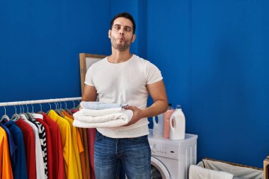 Young hispanic man with beard holding folded clean towels at laundry room making fish face with mouth and squinting eyes, crazy and comical. 