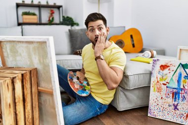 Young man with beard painting canvas at home afraid and shocked, surprise and amazed expression with hands on face 
