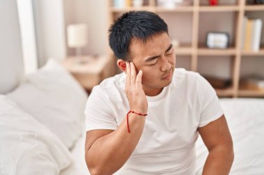 Young chinese man suffering for ear pain sitting on bed at bedroom