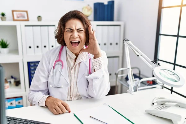Middle age hispanic woman wearing doctor uniform and stethoscope at the clinic shouting and screaming loud to side with hand on mouth. communication concept.