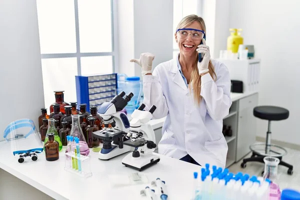 Beautiful woman working at scientist laboratory speaking on the phone pointing thumb up to the side smiling happy with open mouth