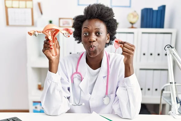 African doctor woman holding anatomical female genital organ and menstrual cup puffing cheeks with funny face. mouth inflated with air, catching air.