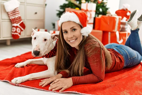 Young caucasian woman smiling confident lying with dog by christmas tree at home