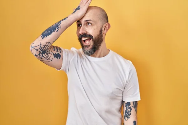 Young hispanic man with tattoos standing over yellow background surprised with hand on head for mistake, remember error. forgot, bad memory concept.