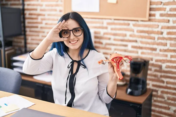 Young girl with blue hair holding model of female genital organ at the office complaining for menstruation pain smiling happy doing ok sign with hand on eye looking through fingers