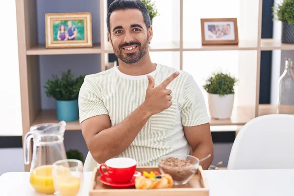 Hispanic man with beard eating breakfast smiling cheerful pointing with hand and finger up to the side
