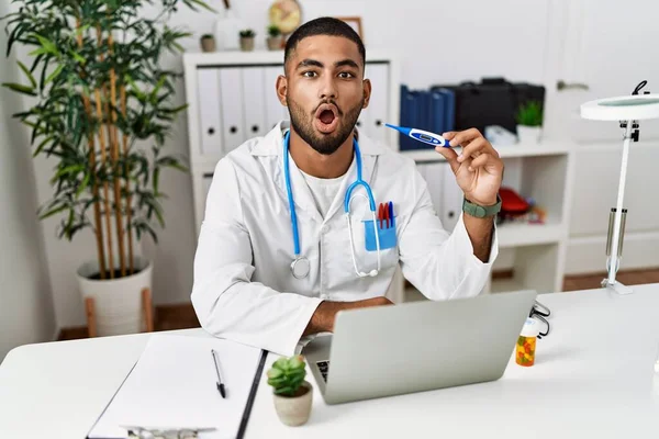 Young indian doctor holding thermometer scared and amazed with open mouth for surprise, disbelief face