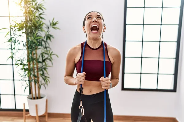 Young hispanic woman training arm resistance with elastic arm bands at sport center angry and mad screaming frustrated and furious, shouting with anger looking up.
