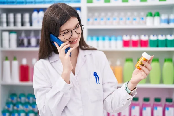 年轻的高加索女药剂师拿着药瓶在药店用智能手机交谈 — 图库照片