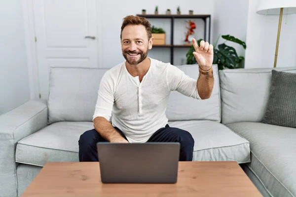 Middle age man using laptop at home smiling and confident gesturing with hand doing small size sign with fingers looking and the camera. measure concept.