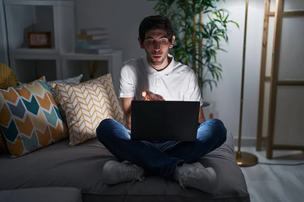 Young hispanic man using laptop at home at night looking at the camera blowing a kiss with hand on air being lovely and sexy. love expression.
