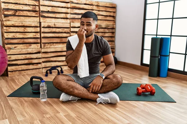 Young indian man sitting on training mat at the gym looking stressed and nervous with hands on mouth biting nails. anxiety problem.
