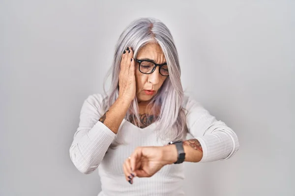 Middle age woman with grey hair standing over white background looking at the watch time worried, afraid of getting late