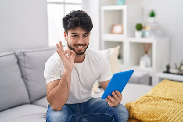 Hispanic man with beard using touchpad sitting on the sofa smiling positive doing ok sign with hand and fingers. successful expression.