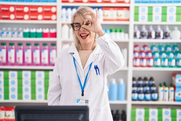 Young caucasian woman working at pharmacy drugstore doing ok gesture with hand smiling, eye looking through fingers with happy face.