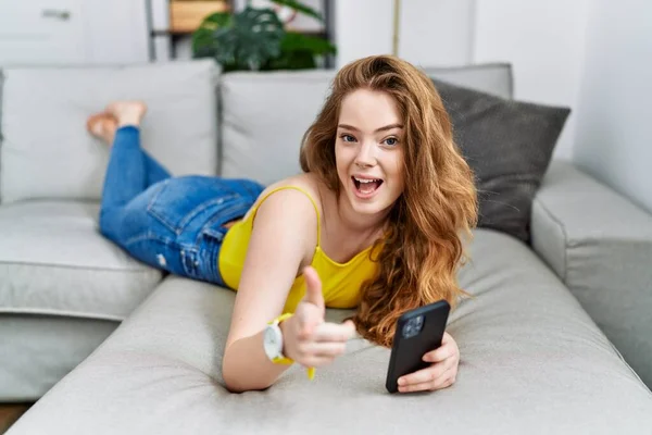 若い白人女性は 幸せと面白い顔を持つカメラに指を指してスマートフォンを使用してソファの上に横たわっていた 良いエネルギーと振動は — ストック写真