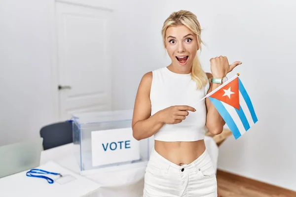 Young caucasian woman at political campaign election holding cuba flag pointing thumb up to the side smiling happy with open mouth