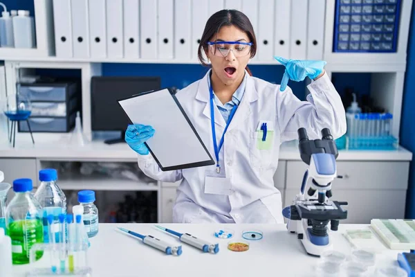 Hispanic young woman working at scientist laboratory pointing down with fingers showing advertisement, surprised face and open mouth