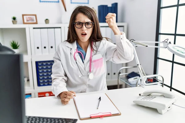 Young doctor woman wearing doctor uniform and stethoscope at the clinic angry and mad raising fist frustrated and furious while shouting with anger. rage and aggressive concept.