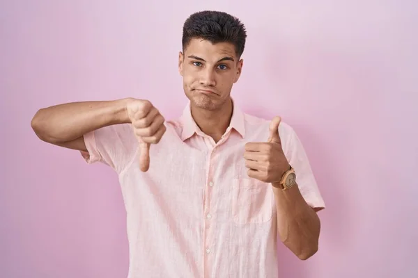 Young hispanic man standing over pink background doing thumbs up and down, disagreement and agreement expression. crazy conflict