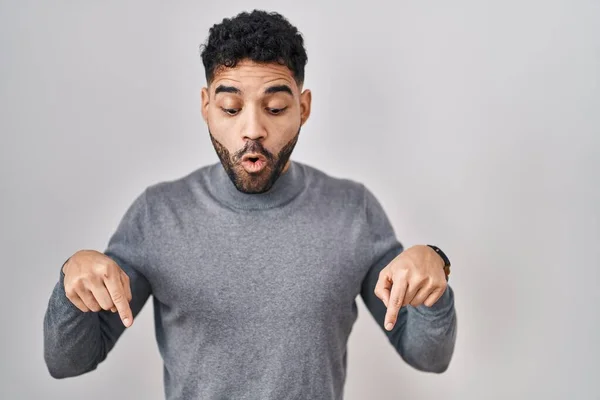 Hispanic man with beard standing over white background pointing down with fingers showing advertisement, surprised face and open mouth