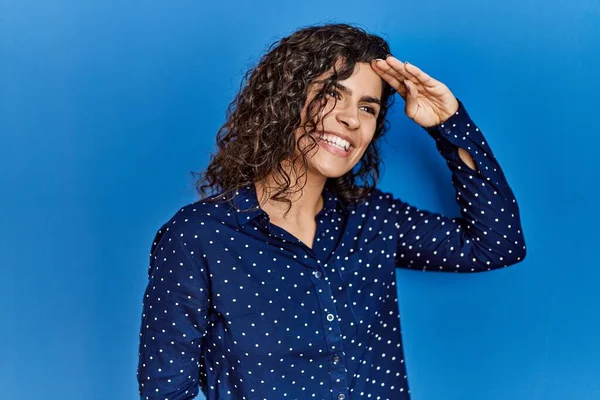 Young brunette woman with curly hair wearing casual clothes over blue background very happy and smiling looking far away with hand over head. searching concept.