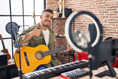 Young hispanic man doing online guitar tutorial smiling happy and positive, thumb up doing excellent and approval sign 