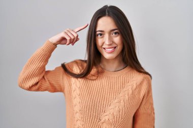 Beyaz arka planda duran esmer kadın gülümseyerek tek parmağıyla kafasını işaret ediyor, harika bir fikir ya da düşünce, iyi bir hafıza. 