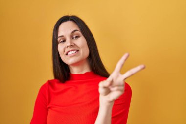 Sarı arka planda durup gülümseyen genç İspanyol kadın parmaklarını zafer işareti yaparken gösteren kameraya bakıyor. İki numara.. 