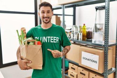 Young hispanic man wearing volunteer t shirt holding groceries smiling happy pointing with hand and finger 