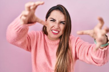 Young hispanic woman standing over pink background shouting frustrated with rage, hands trying to strangle, yelling mad 