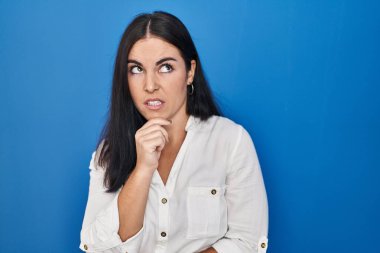 Mavi arka planda dikilen genç İspanyol kadın bir soru hakkında endişeli ve gergin bir şekilde düşünüyor. 