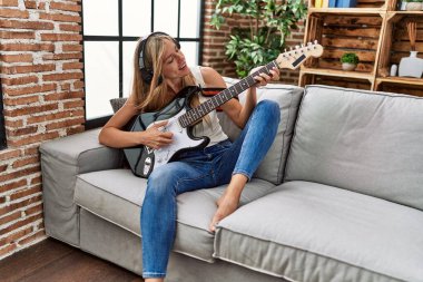 Genç sarışın kadın evde gitar çalarken kendinden emin gülümsüyor.