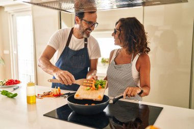 Orta yaşlı İspanyol çift, gülümsüyor. Mutfakta tavaya yiyecek koyuyorlar.