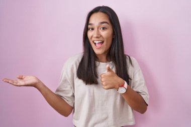 Pembe arka planda duran genç İspanyol kadın avuç içini gösteriyor ve baş parmaklarıyla iyi bir hareket yapıyor, mutlu ve neşeli gülümsüyor. 